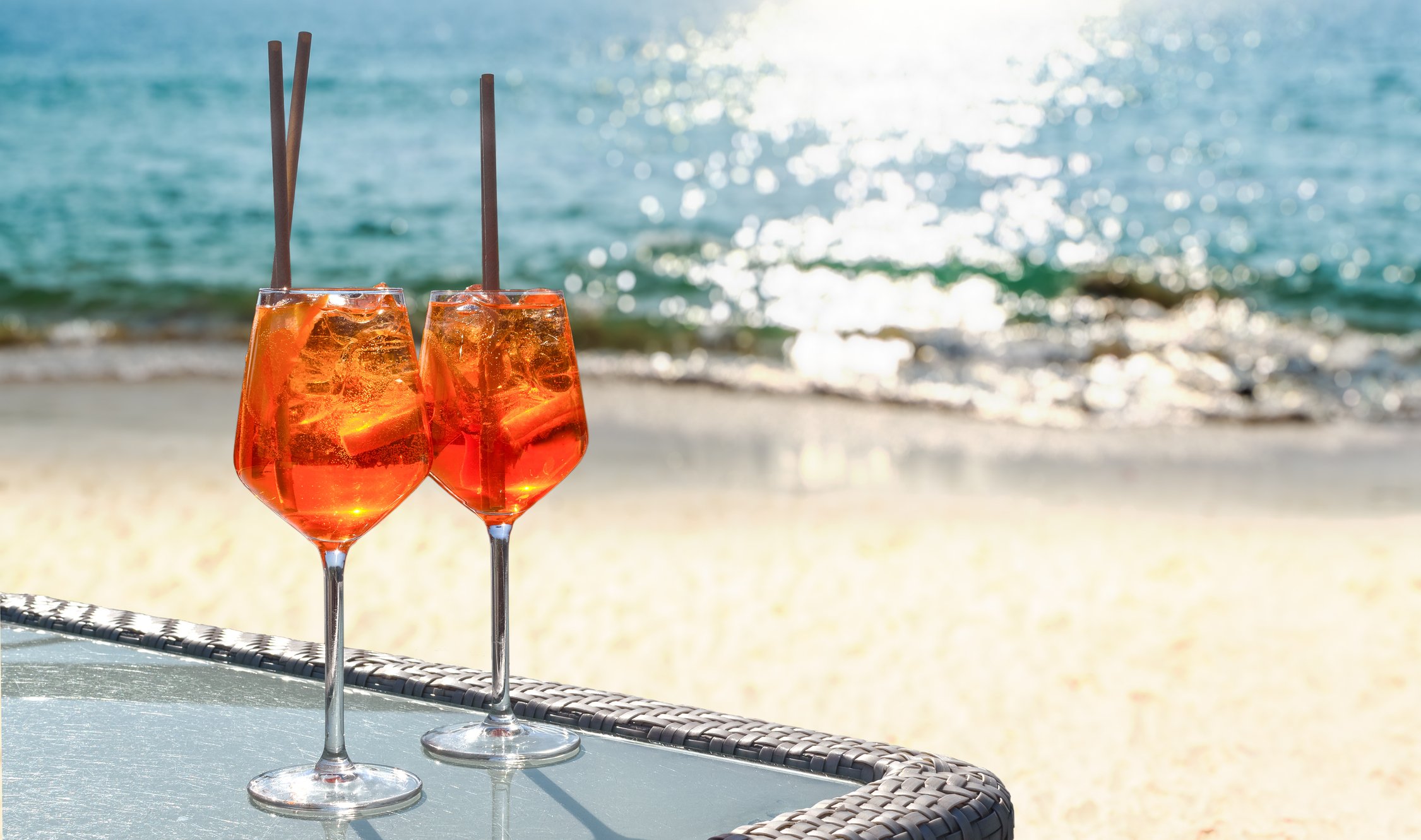 Duas taças de Aperol Spritz com canudos posicionadas em uma mesa de praia, com o mar azul e areia clara ao fundo.