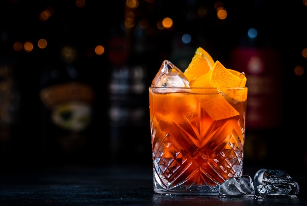 Close de um copo de Aperol Negroni com fatias de laranja e gelo, destacando a cor alaranjada vibrante contra um fundo escuro e desfocado.