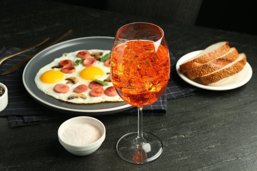 Uma taça de Aperol Spritz em primeiro plano ao lado de um prato de café da manhã com ovos fritos, bacon e pães.