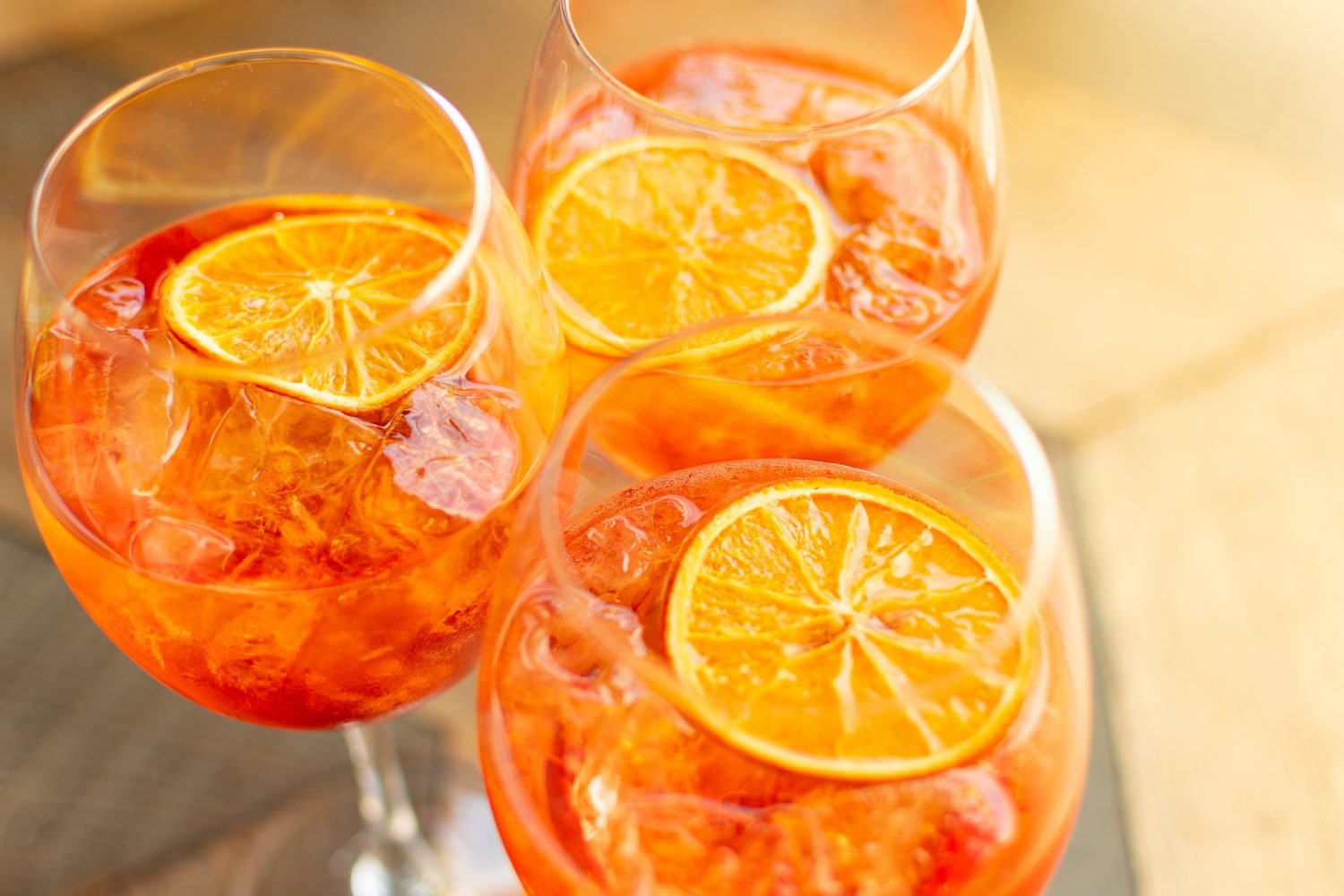 Vista de cima de três taças de Aperol Spritz com gelo e rodelas de laranja flutuando, dispostas em uma superfície rústica.