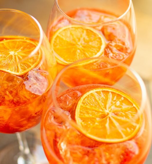 Vista de cima de três taças de Aperol Spritz com gelo e rodelas de laranja flutuando, dispostas em uma superfície rústica.