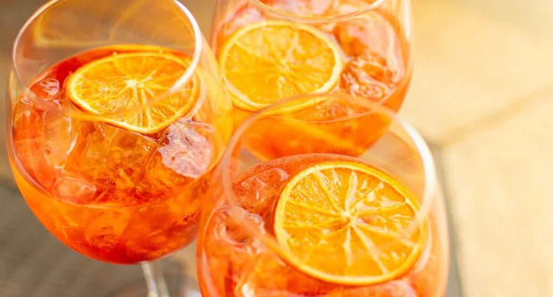 Vista de cima de três taças de Aperol Spritz com gelo e rodelas de laranja flutuando, dispostas em uma superfície rústica.