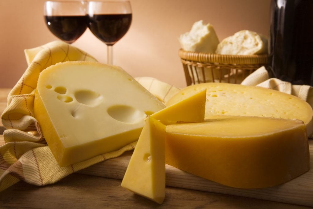 Fatias de queijo em uma mesa com taças de vinho tinto e cesto de pão ao fundo