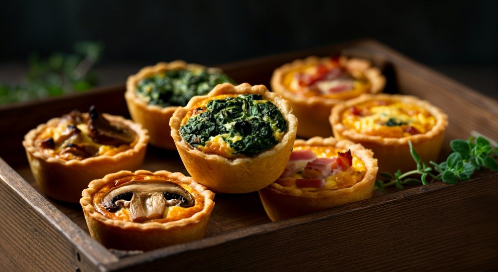 Quiches individuais em massa crocante com recheios de espinafre e queijo, apresentadas em tábua de madeira escura - prato elegante e prático para piquenique