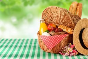 Cesta de vime com frutas vermelhas, pão francês, suco natural e chapéu de palha sobre toalha xadrez verde e branca para piquenique