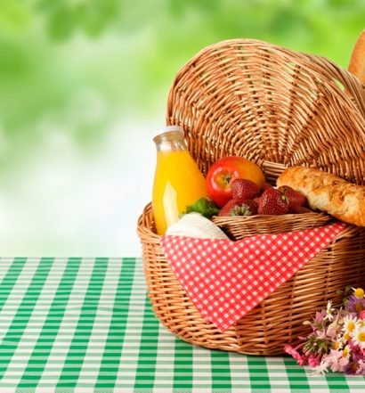 Cesta de vime com frutas vermelhas, pão francês, suco natural e chapéu de palha sobre toalha xadrez verde e branca para piquenique