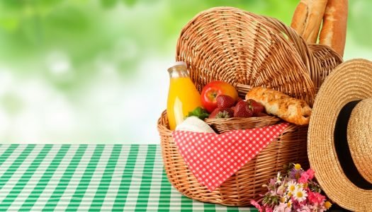 Cesta de vime com frutas vermelhas, pão francês, suco natural e chapéu de palha sobre toalha xadrez verde e branca para piquenique