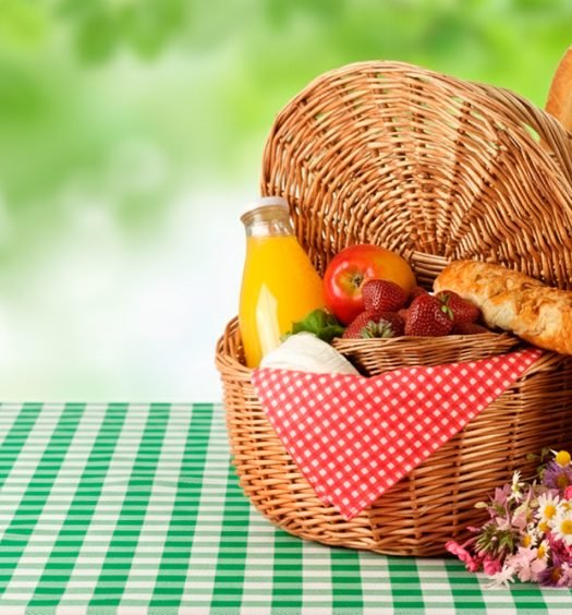 Cesta de vime com frutas vermelhas, pão francês, suco natural e chapéu de palha sobre toalha xadrez verde e branca para piquenique