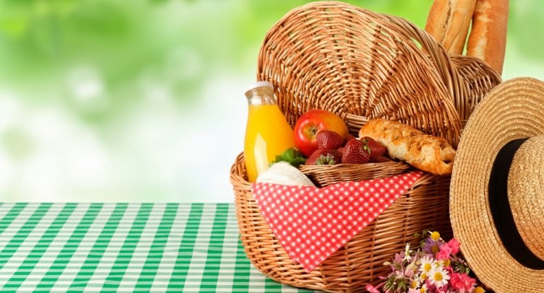 Cesta de vime com frutas vermelhas, pão francês, suco natural e chapéu de palha sobre toalha xadrez verde e branca para piquenique