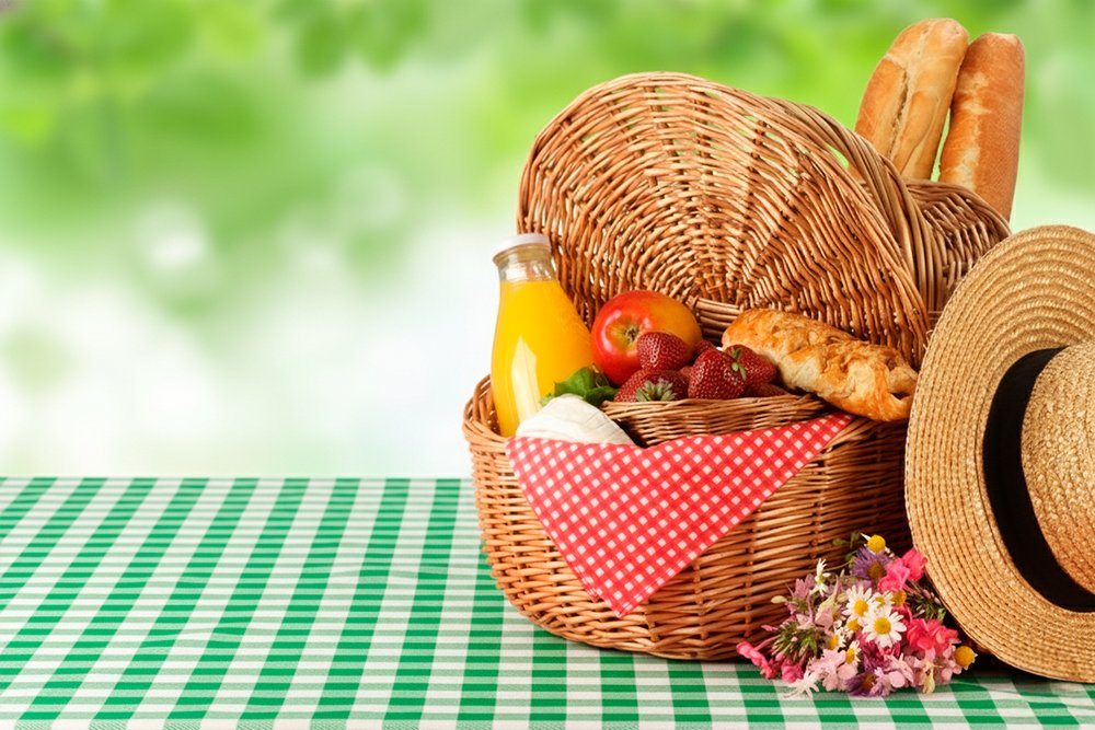 Cesta de vime com frutas vermelhas, pão francês, suco natural e chapéu de palha sobre toalha xadrez verde e branca para piquenique
