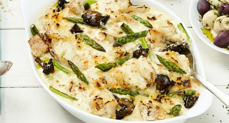 Gratinado de bacalhau com aspargos e cogumelos em molho branco cremoso, servido em travessa oval branca.