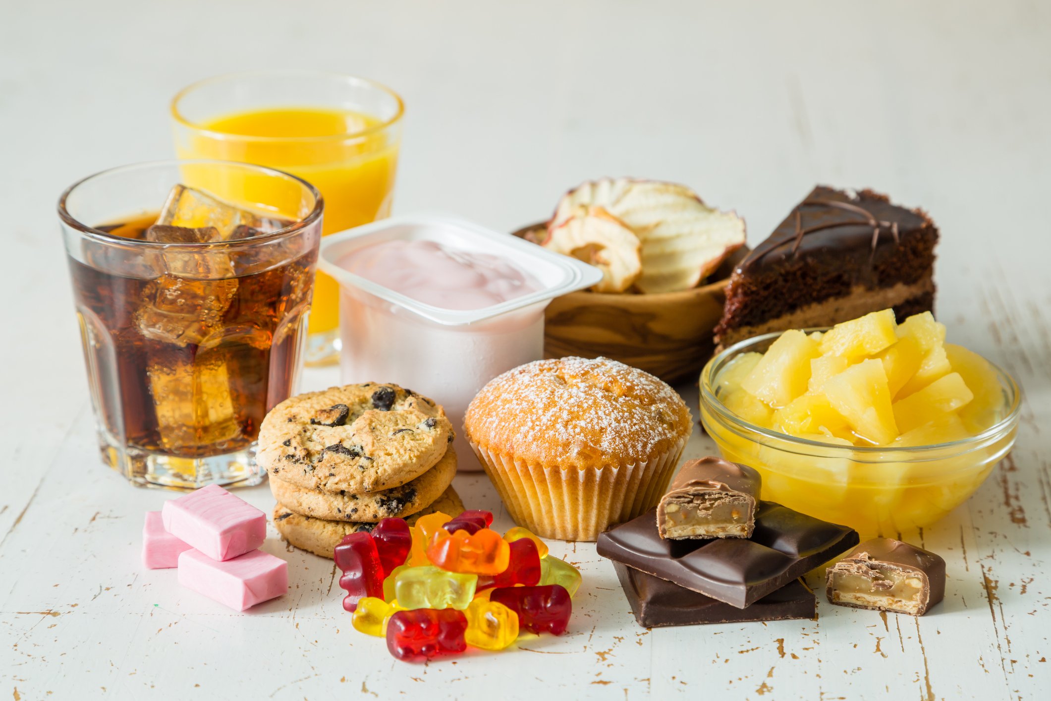Grupo de doces e açúcares na pirâmide alimentar: refrigerante, suco industrializado, iogurte adoçado, bolo, biscoitos, muffins, balas de gelatina, chocolate e abacaxi em calda sobre mesa clara