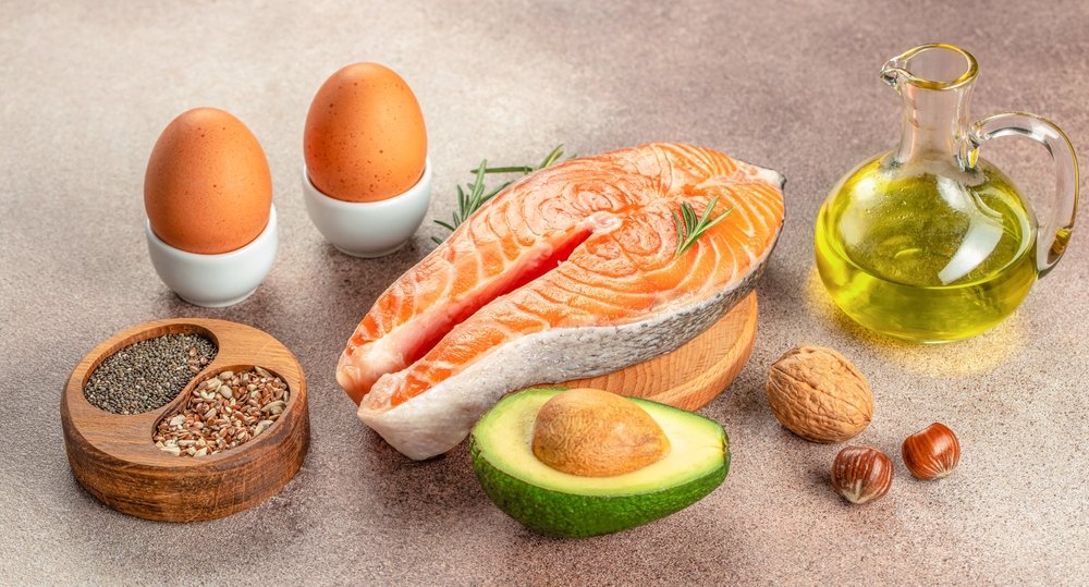 Grupo de gorduras e óleos saudáveis na pirâmide alimentar: ovos, salmão fresco, abacate, azeite de oliva, castanhas e sementes sobre superfície neutra