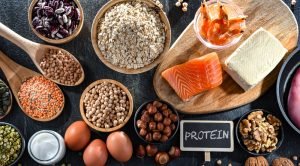 Fontes de proteína da pirâmide alimentar — salmão, tofu, camarão, ovos, feijões, lentilhas, grão-de-bico, aveia e nozes sobre superfície escura.