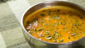 Caldo de sururu com ervas verdes em tigela metálica sobre toalha de mesa texturizada.