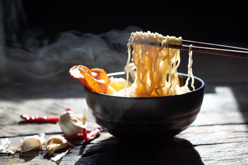 lamen fumegante com noodles levantados por hashis, alho e pimenta ao lado