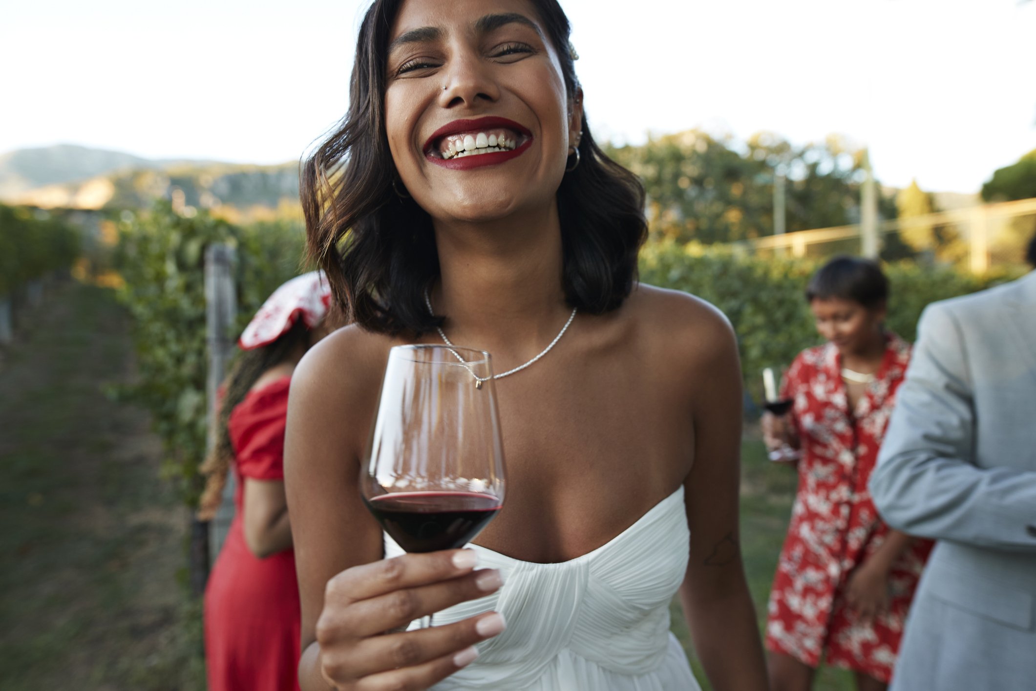 Pessoa sorrindo com taça de vinho tinto em vinhedo; consumo moderado associado à redução do estresse e melhor humor.
