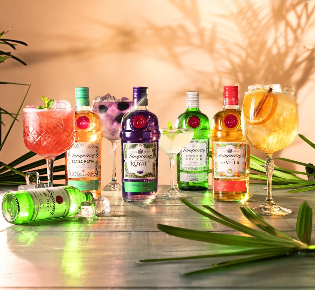 Garrafas de gin Tanqueray com copos de drinks decorados com frutas e ervas em uma mesa iluminada, indicando drinks para o verão