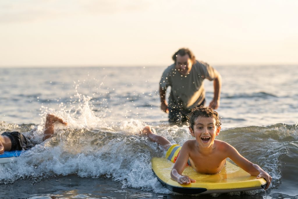 Criança surfando em uma prancha na praia com um adulto ao fundo