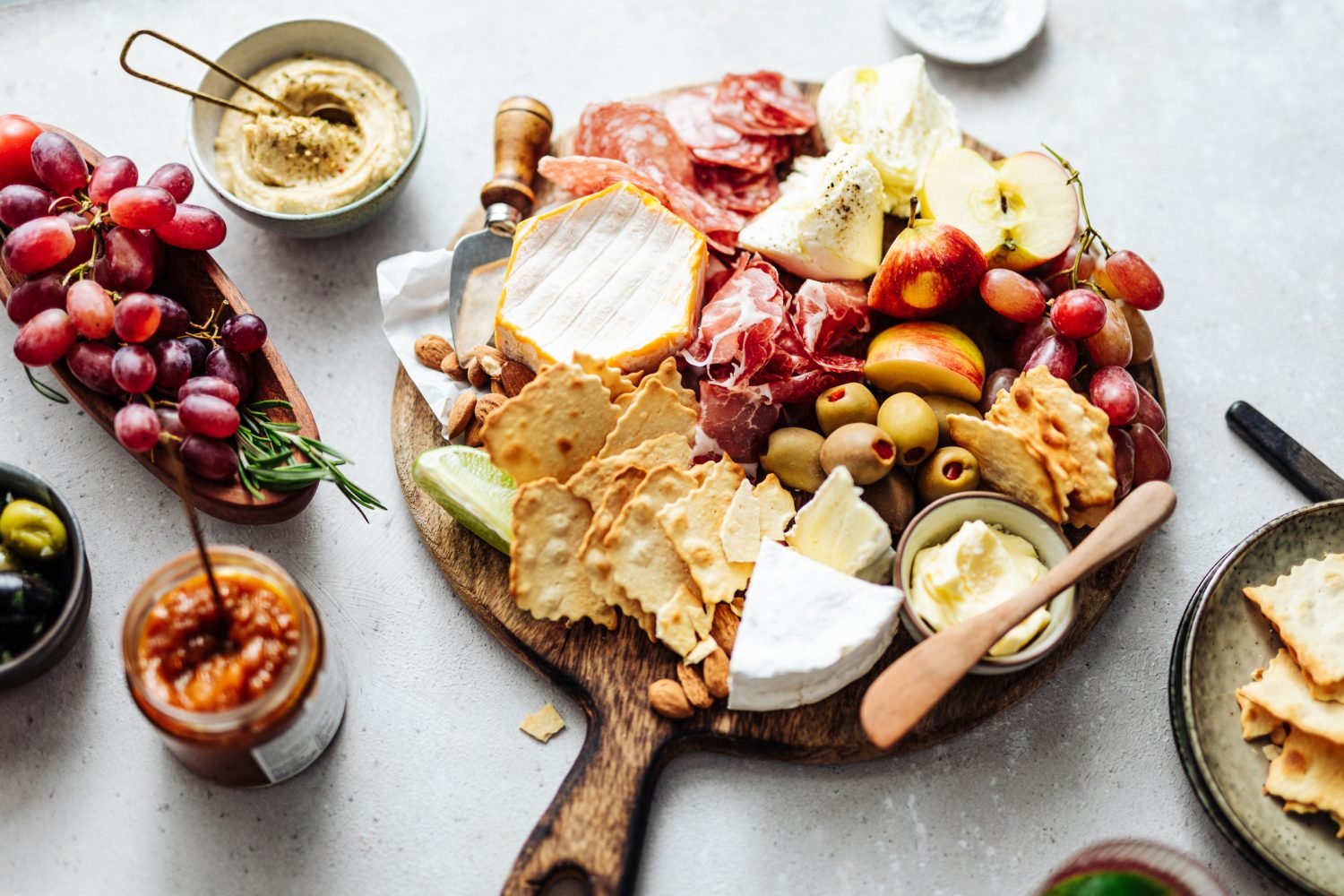 Uma tábua de frios com queijos, uvas, fatias de maçã, biscoitos e acompanhamentos em uma superfície clara