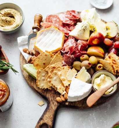 Uma tábua de frios com queijos, uvas, fatias de maçã, biscoitos e acompanhamentos em uma superfície clara