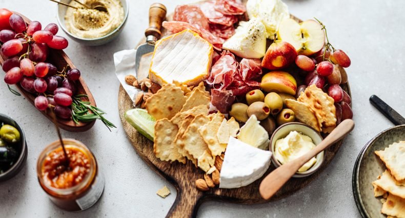 Uma tábua de frios com queijos, uvas, fatias de maçã, biscoitos e acompanhamentos em uma superfície clara