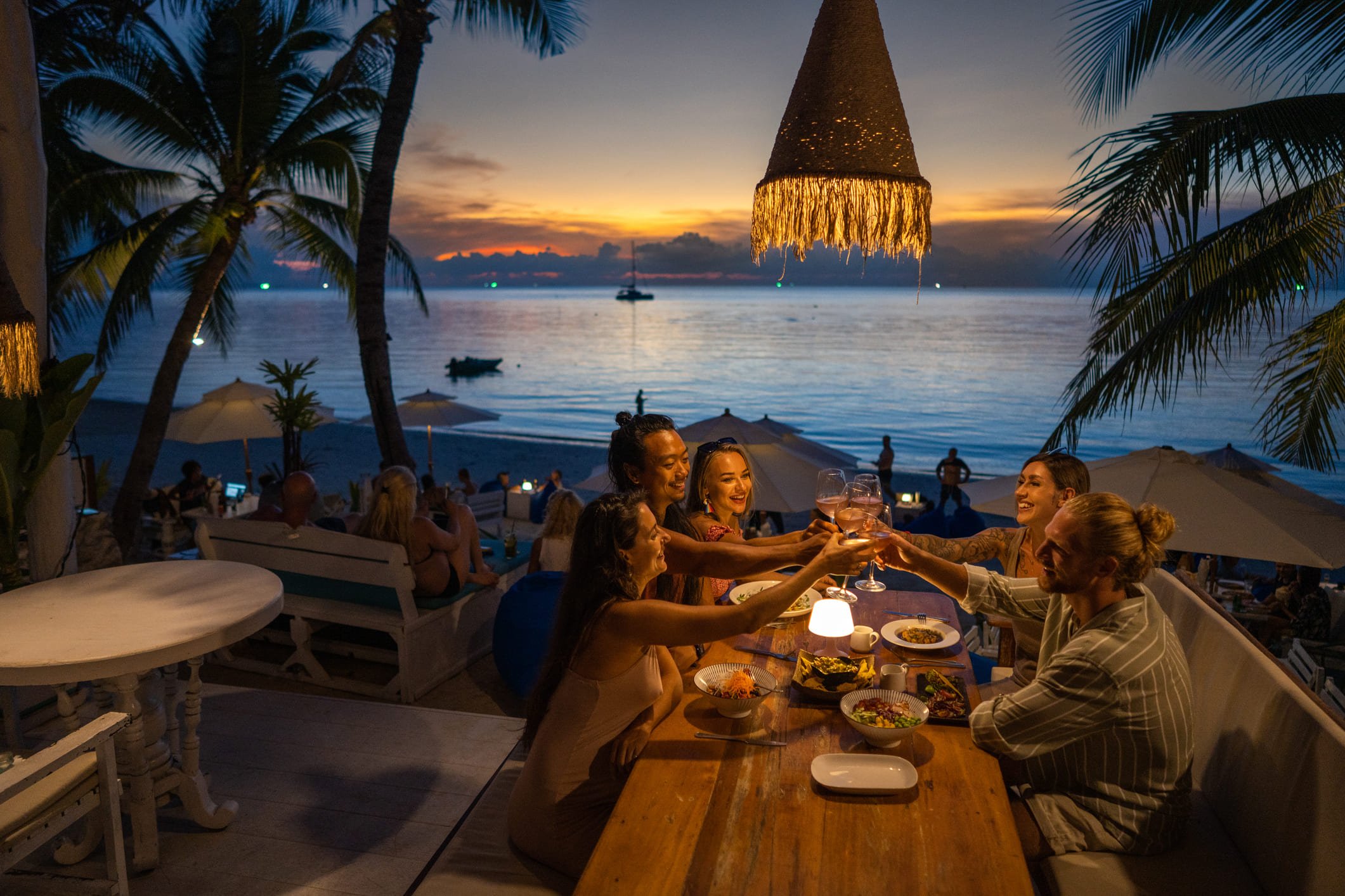 Grupo de amigos levantando copos em um restaurante à beira-mar ao pôr do sol.