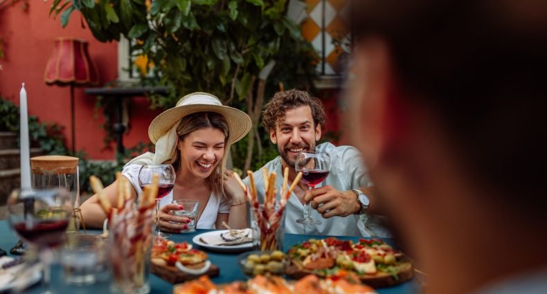 Casal se divertindo em um jantar ao redor de uma mesa cheia de pratos e vinhos.