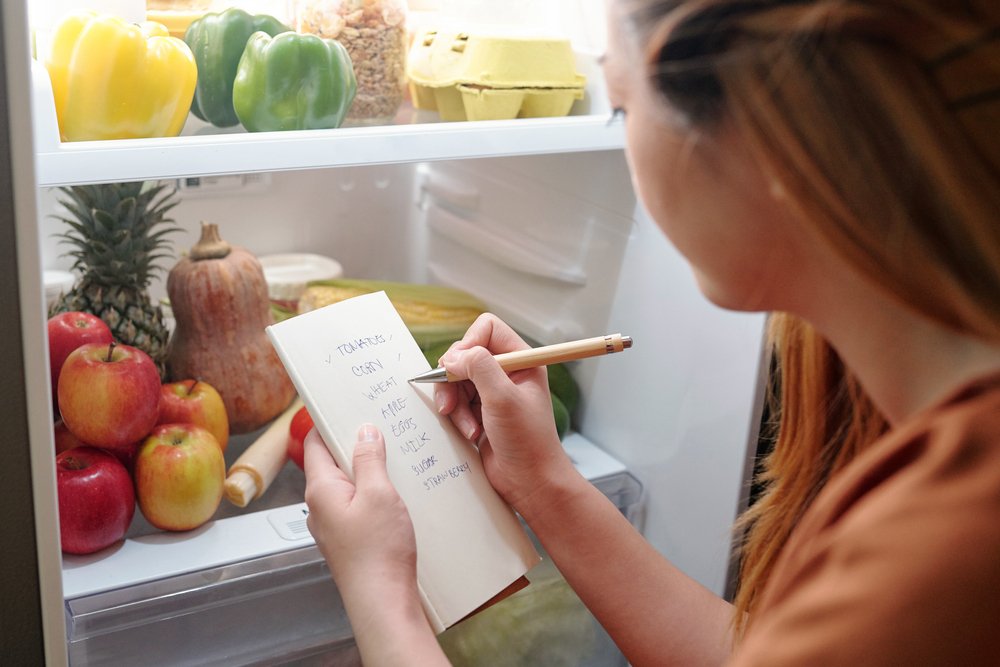 Mulher escrevendo na lista de compras diante da geladeira