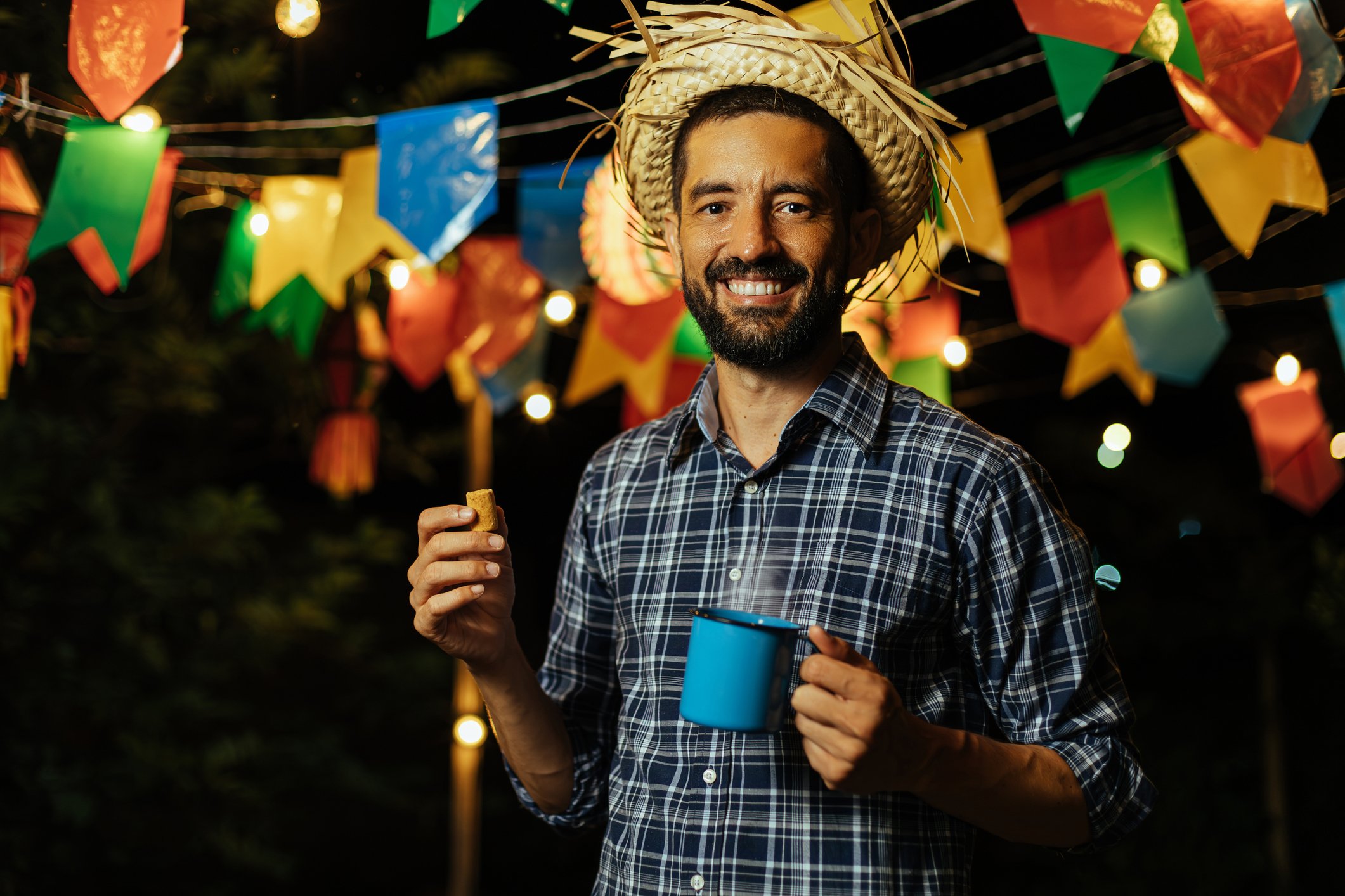 Homem sorridente em festa junina com chapéu de palha e caneca azul.