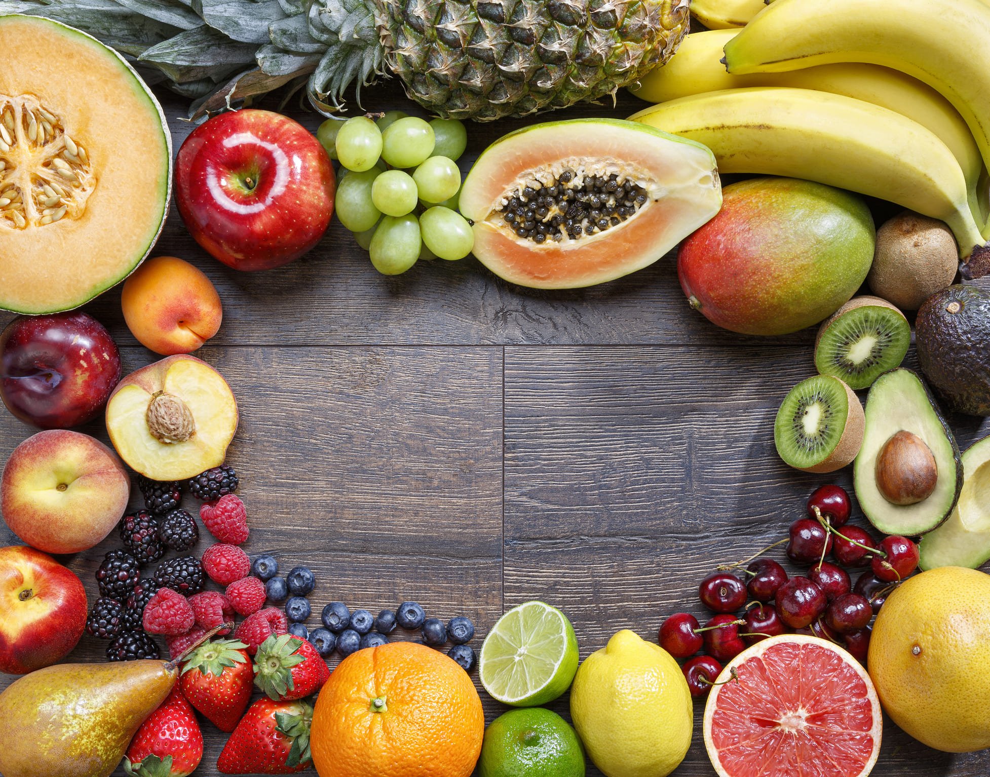 Uma variedade colorida de frutas, incluindo abacaxi, mamão, maçã e uvas, dispostas em uma mesa de madeira.