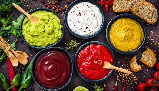 Seis molhos coloridos em tigelas com pão e ervas ao fundo.