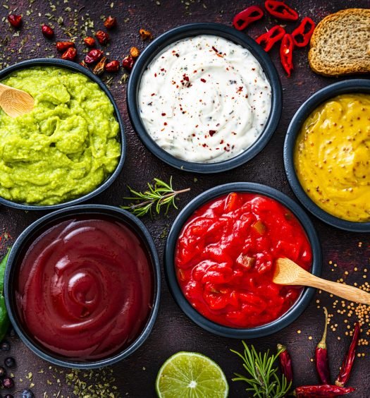 Seis molhos coloridos em tigelas com pão e ervas ao fundo.