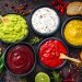 Seis molhos coloridos em tigelas com pão e ervas ao fundo.