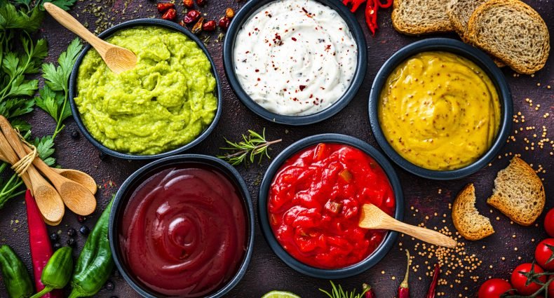 Seis molhos coloridos em tigelas com pão e ervas ao fundo.