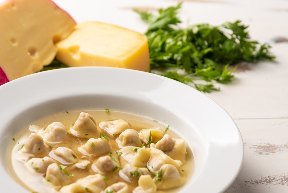 Sopa de capeletti servida em um prato branco com queijo e salsinha ao fundo