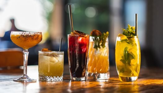 Cinco diferentes copos e taças apresentados com cocktails variados em um ambiente iluminado