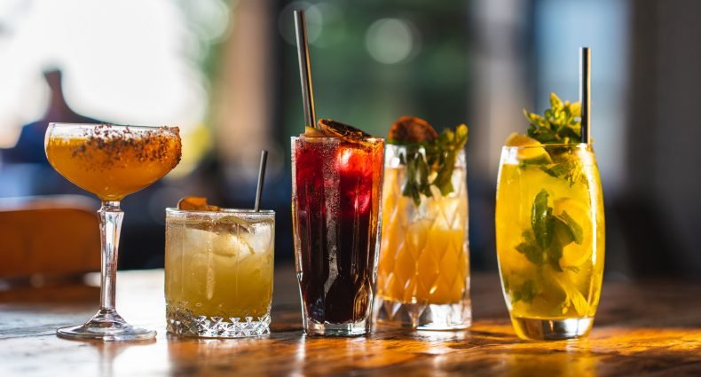 Cinco diferentes copos e taças apresentados com cocktails variados em um ambiente iluminado