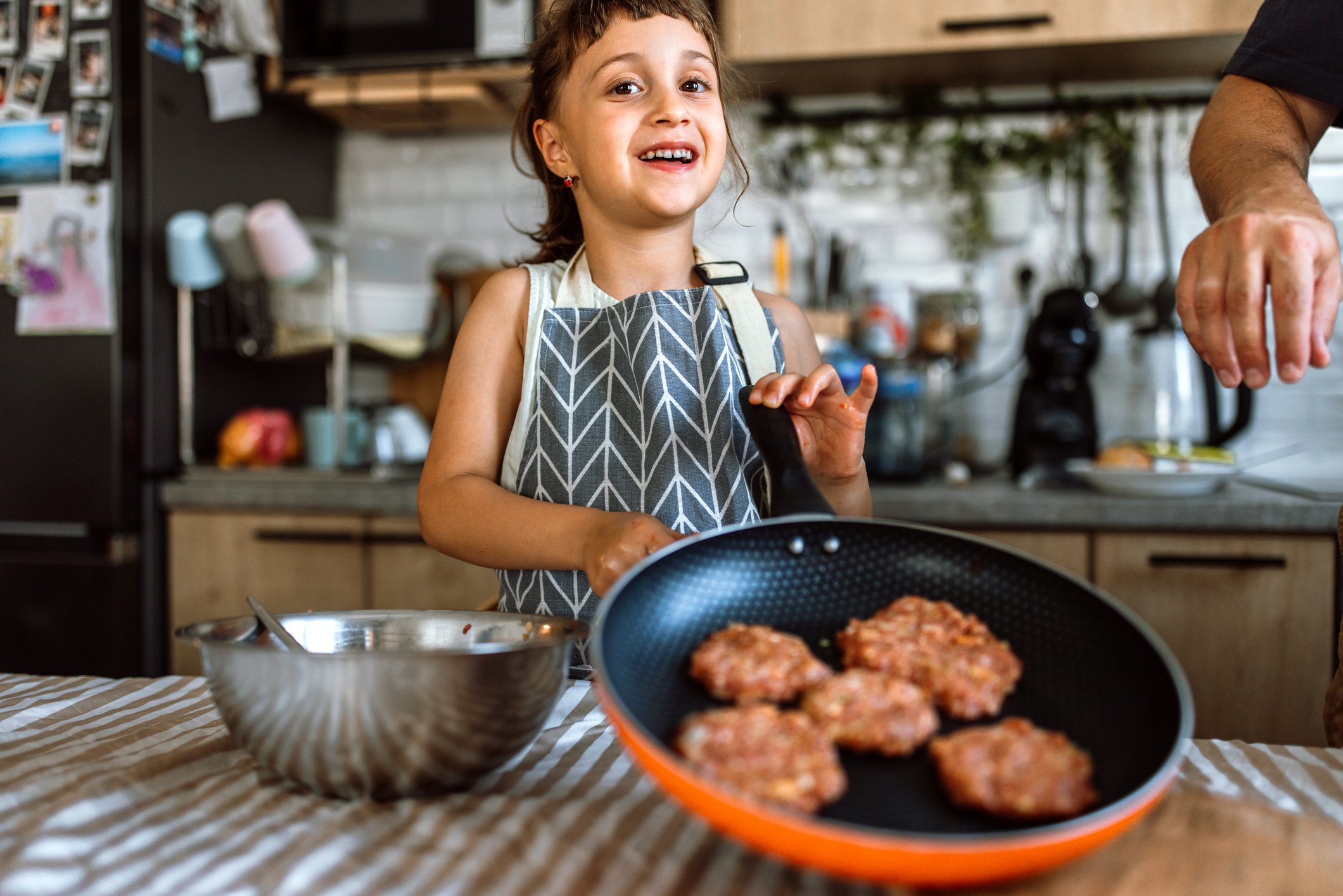 Menina concentrada preparando hambúrgueres ou panquecas em uma frigideira, usando avental e cozinhando com segurança.