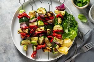 Espetinhos veganos com abobrinha grelhada, pimentão vermelho, tomate-cereja e cebola-roxa em espetos de metal — refeição vegana saudável.