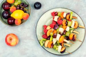 Espetinhos de frutas variadas (kiwi, banana, uva, framboesa, ameixa, damasco) em um prato, com um bowl de frutas frescas ao lado, ideais para uma sobremesa saudável e divertida para crianças.
