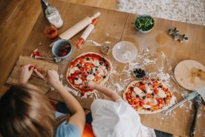 Vista aérea de duas pessoas (adulto e criança) fazendo pizzas caseiras com tomate, azeitona e queijo em uma mesa de cozinha com farinha e utensílios.