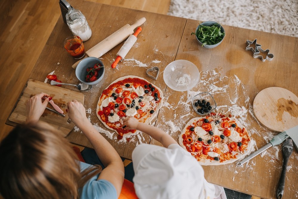 Vista aérea de duas pessoas (adulto e criança) fazendo pizzas caseiras com tomate, azeitona e queijo em uma mesa de cozinha com farinha e utensílios.