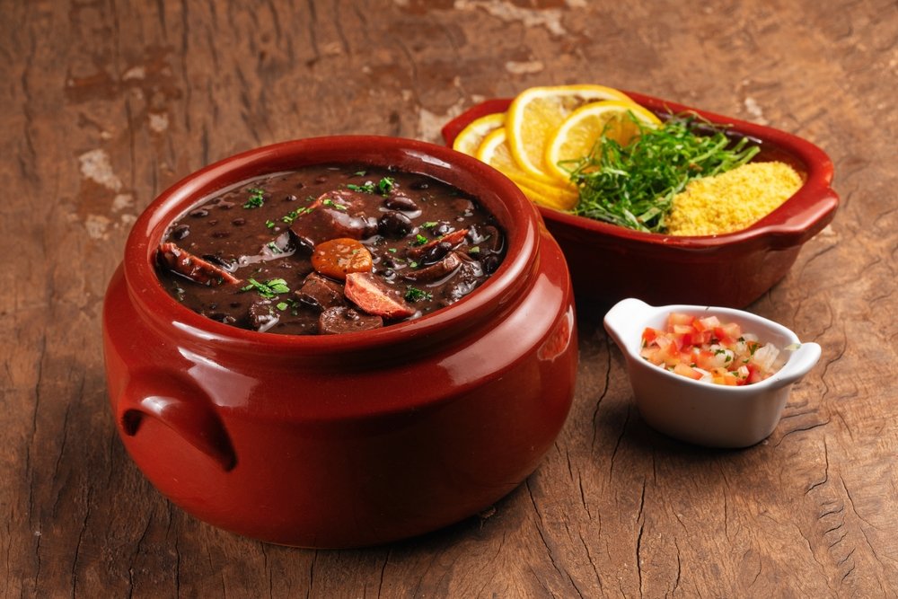 Feijoada vegana servida em tigela vermelha com acompanhamentos: polenta, broa de milho, fatias de laranja e molho de pimenta caseiro