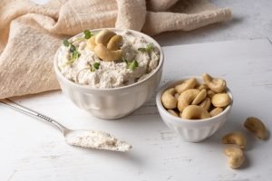 Creme de queijo vegano feito de castanha-de-caju em tigela branca ao lado de castanhas inteiras.