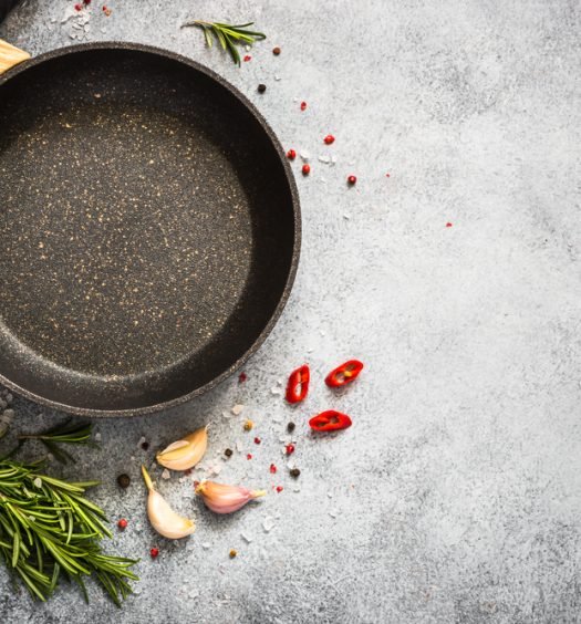 Panela antiaderente vazia com cabo de madeira vista de cima sobre fundo cinza, rodeada de alecrim, alho e pimenta