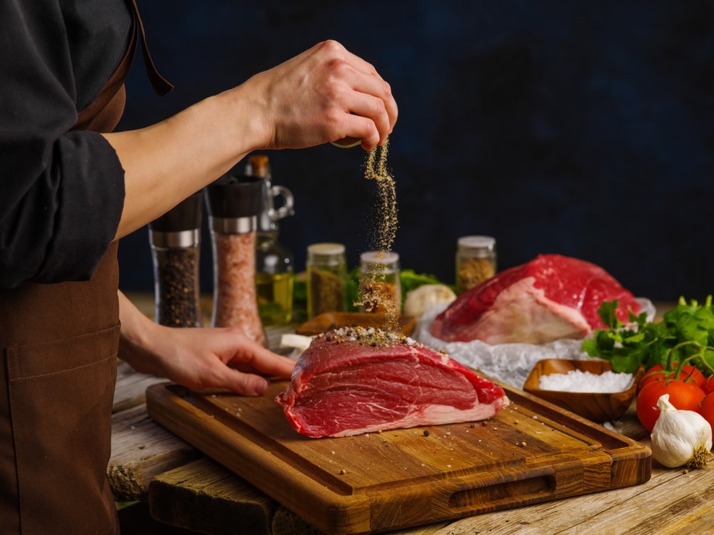 Mão polvilhando tempero em pó sobre filé de carne em tábua de corte, com potes de especiarias, tomate e alho ao fundo, técnica de preparo profissional