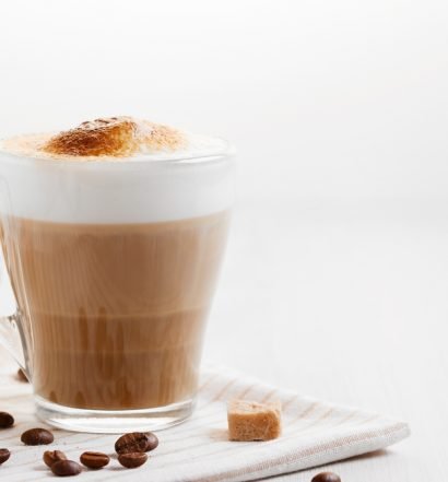 Cappuccino cremoso em xícara de vidro com espuma alta, grãos de café e cubos de açúcar mascavo sobre pano listrado