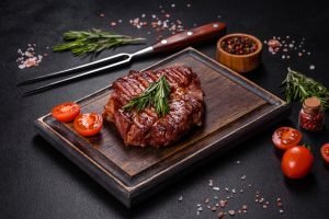 Carne vermelha grelhada com rosmaninho, tomate fresco e pimenta do reino em apresentação gourmet em tábua de madeira, com temperos em especiaria