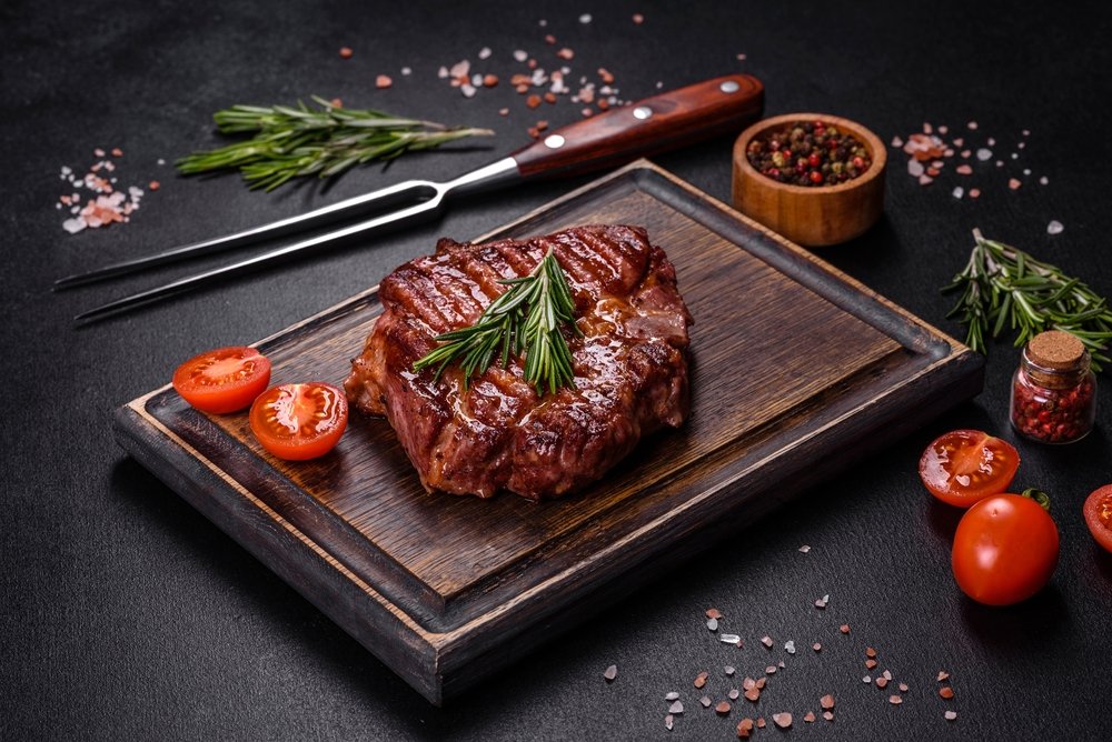 Carne vermelha grelhada com rosmaninho, tomate fresco e pimenta do reino em apresentação gourmet em tábua de madeira, com temperos em especiaria